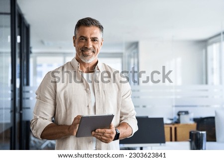 Latin Hispanic mature adult professional business man looking at camera and smiling. Indian senior businessman CEO holding digital tablet using fintech tab application standing inside company office. Royalty-Free Stock Photo #2303623841