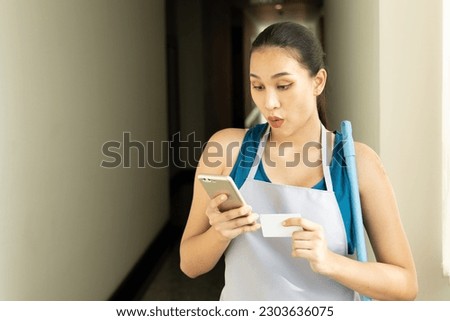 Excited positive Asian cleaning service worker woman, using smartphone with good connection Royalty-Free Stock Photo #2303636075