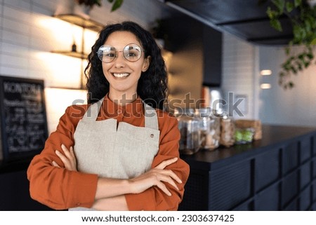 Portrait of young beautiful woman worker in cafe restaurant, Hispanic woman with curly hair and glasses smiling and looking at camera with crossed arms and wearing an apron.