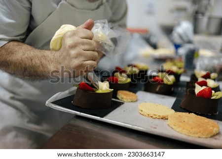 Close-up picture of the hands of a senior pastry chef decorating cakes with fruits with a pastry bag in a bakery kitchen