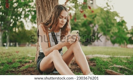 Beautiful girl with long wavy hair wearing in white top uses mobile phone while sitting on the green grass under tree in the park. Girl flip through the pictures on your phone with fingers