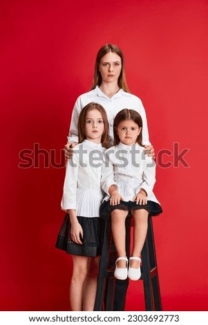 Portrait of beautiful young woman and two cute little girls in formal wear posing against red studio background. Serious expression. Concept of family, motherhood, childhood, fashion, lifestyle
