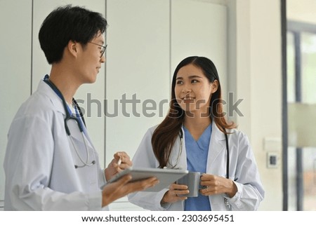 Two professional doctors discuss patient diagnosis data on digital tablet, standing in medical office
