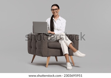 Happy woman with laptop sitting in armchair on light gray background