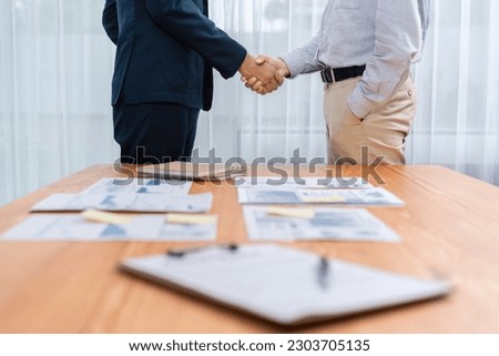 Two businesspeople shake hand after signing contract document to merge their partnerships in conference room and finalized pile of papers of financial report and data analysis on meeting table. Entity Royalty-Free Stock Photo #2303705135