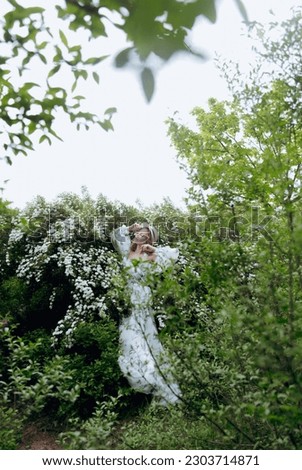 Beautiful, stylish, cute blonde in a white dress with a green pattern as a model for a photo shoot in the bush bride, spirea. They made luxurious portraits of her in the green garden of the park.