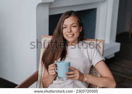 Cheerful hispanic young woman in white t-shirt sitting on chair holds cup of coffee looks at camera toothy smiles enjoying vacation home, hotel room during holidays. Successful girl relaxing.