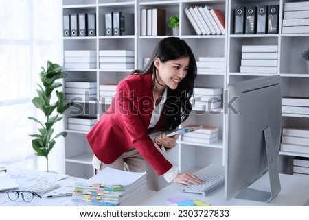 Young business smiling happy asian woman working holding a piece of paperwork and reading while standing in front of laptop at home, work from home concept