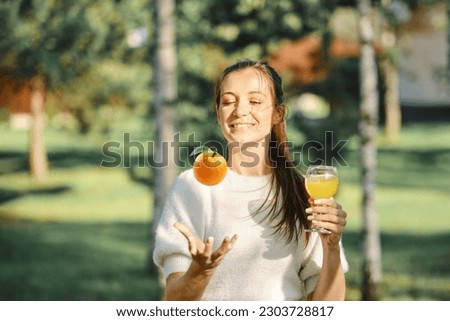 Joyful woman tossing orange with one hand while holding glass with fresh juice in another hand Royalty-Free Stock Photo #2303728817