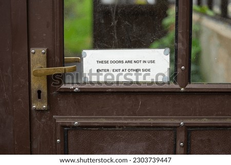 Small laminated printed white paper sign tacked with grey sticky paste to window glass pane of brown metal door with bronze handle warning "THESE DOORS ARE NOT IN USE ENTER EXIT AT OTHER SIDE" 