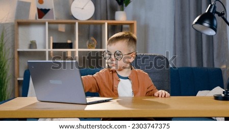 Front view of handsome small interested kid in round eyeglasses which sitting in front of computer and revisioning cartoon or playing games
