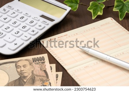 Passbook, calculator and Japanese money. The passbook is written in Japanese as "ordinary savings", "date", "handling store", "deposit amount", "payment amount", and "current balance". Royalty-Free Stock Photo #2303754623
