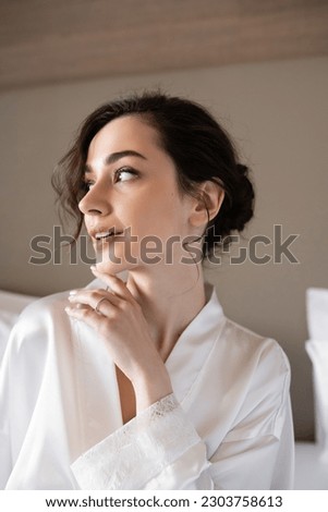 young woman with brunette hair in white silk robe showing engagement ring on finger while looking away in hotel room on wedding day, special occasion, young bride