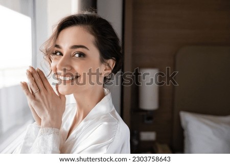 portrait of delightful young woman with engagement ring on finger standing in white silk robe and looking at camera in modern hotel suite, special occasion, bride on wedding day