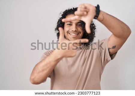 Hispanic man with curly hair standing over white background smiling making frame with hands and fingers with happy face. creativity and photography concept. 