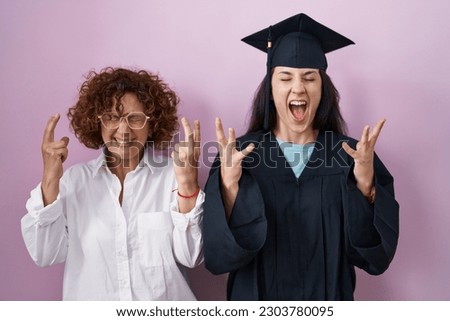 Hispanic mother and daughter wearing graduation cap and ceremony robe celebrating mad and crazy for success with arms raised and closed eyes screaming excited. winner concept 