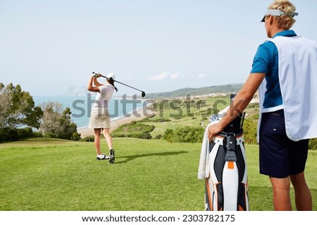Caddy watching woman tee off on golf course overlooking ocean Royalty-Free Stock Photo #2303782175