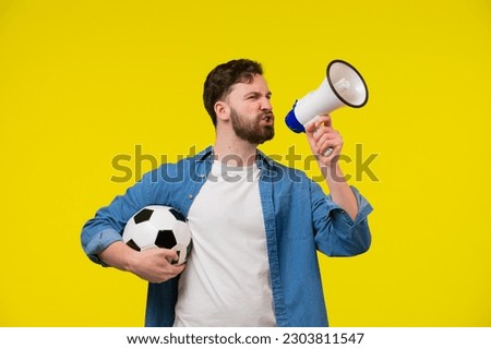 Very happy cheerful man with beard in striped t-shirt blowing in big bullhorn holding football ball, celebrating championship beginning. Indoor studio shot isolated on yellow background