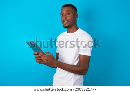 Photo of optimistic young man wearing white T-shirt over blue studio background hold tablet