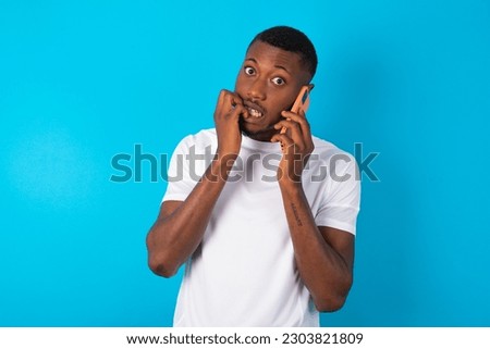 Afraid funny young man wearing white T-shirt over blue studio background holding telephone and bitting nails