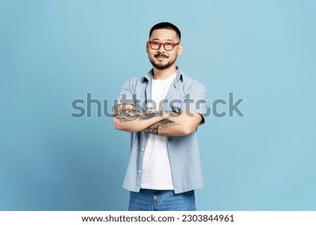 Smiling Asian man wearing stylish red eyeglasses and casual clothes isolated on blue background. Portrait of successful asian freelancer  with crossed arms looking at camera