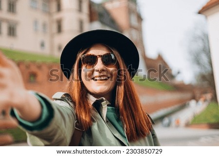 Attractive young Redhead female tourist is exploring new city. Traveling. Happy optimistic girl walking in city and makes selfie poses against historic building in Krakow.