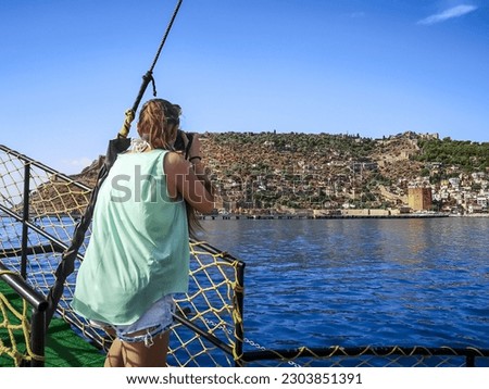 Young female photographer takes pictures of Red Tower in Alanya (Turkey) while on the deck of a ship. Woman with a camera in the Mediterranean sea on the background of the peninsula with the old town