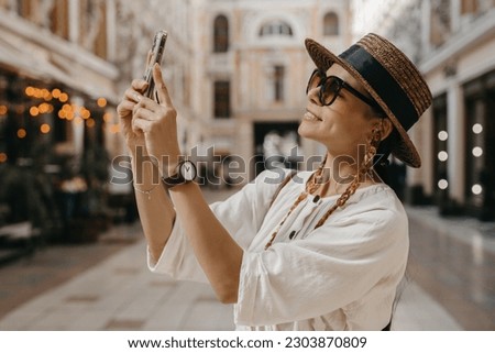 attractive woman walking in shopping street in Italy on vacation dressed in white summer fashion dress straw hat and purse, smiling happy having fun, taking pictures on smartphone