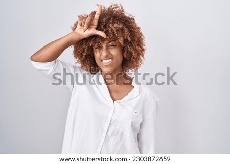 Young hispanic woman with curly hair standing over white background making fun of people with fingers on forehead doing loser gesture mocking and insulting. 