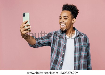 Young smiling happy man of African American ethnicity he wear blue shirt doing selfie shot on mobile cell phone isolated on plain pastel light pink background studio portrait. People lifestyle concept