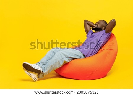 Full body young man of African American ethnicity he wear casual clothes purple t-shirt sit in bag chair hold hands behind neck isolated on plain yellow background studio portrait. Lifestyle concept