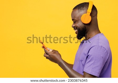 Side view young man of African American ethnicity wear casual clothes purple t-shirt headphones listen to music hold use mobile cell phone isolated on plain yellow background studio. Lifestyle concept Royalty-Free Stock Photo #2303876345