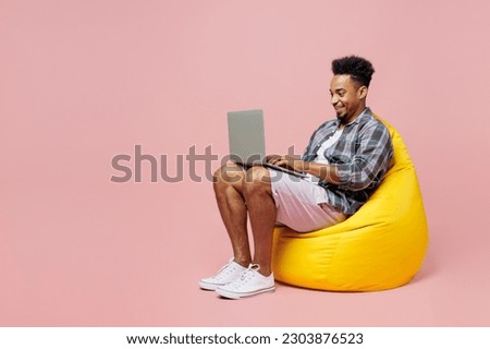 Full body smiling young man of African American ethnicity wear blue shirt sit in bag chair hold use work on laptop pc computer isolated on plain pastel light pink background. People lifestyle concept