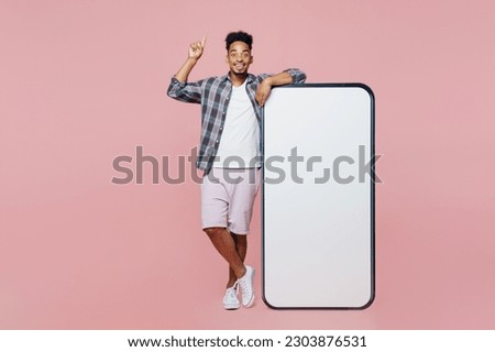 Full body young man of African American ethnicity wear blue shirt stand near big huge blank screen mobile cell phone copy space point finger up new idea isolated on plain pastel light pink background