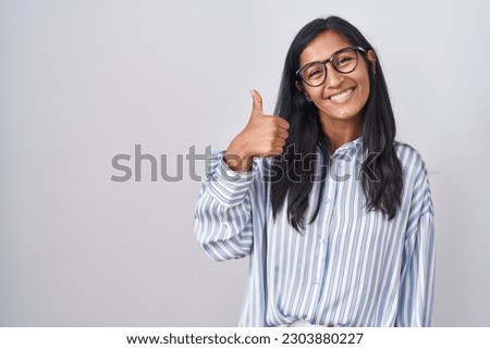 Young hispanic woman wearing glasses doing happy thumbs up gesture with hand. approving expression looking at the camera showing success. 