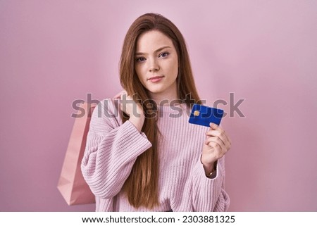 Young caucasian woman holding shopping bag and credit card relaxed with serious expression on face. simple and natural looking at the camera. 