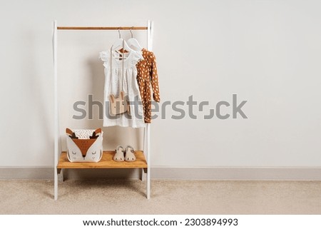 Clothing Rack with kids outfits and storage baskets in children's room. Fashion clothes in white, beige and brown colors on hangers in wardrobe. Set of kids clothes and accessories.   Royalty-Free Stock Photo #2303894993