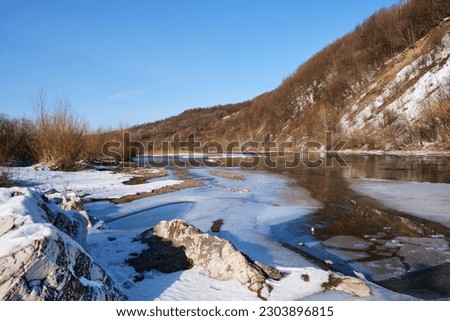Winter landscape on a clear day. A view of a mountain river with ice and mountains covered with snow and stones with different structures and a clear sky