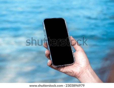 Mobile beach phone with empty screen. Mockup image of black blank desktop screen in hand holding mobile smart phone on the blue sea background in summer. Travel booking, searching, business marketing.