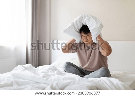 Angry sleepless nervous middle aged asian man with closed eyes sitting in bed and covering ears, head with pillow, hearing and suffering from too loud sound, tired of noisy neighbors, copy space Royalty-Free Stock Photo #2303906377