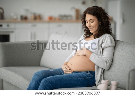 Pregant woman gently caressing her baby bump while relaxing on couch at home, beautiful young expectant mother tenderly touching belly and smiing, enjoying pregnancy time, copy space Royalty-Free Stock Photo #2303906397