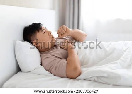 Unhealthy sick middle aged asian man smoker wearing pajamas lying in bed and coughing, touching his chest, home interior, side view, copy space. Lung cancer, pneumonia, smokers morning cough Royalty-Free Stock Photo #2303906429