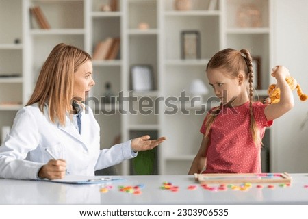 Aggressive Child Therapy. Cute Little Girl Feeling Angry During Therapy Session With Psychotherapist Lady, Upset Small Kid Threaten Specialist Doctor With A Toy, Having Temper Tantrum Royalty-Free Stock Photo #2303906535