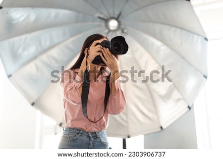 Studio magic. Young photographer woman capturing moments with professional camera in front of reflective umbrella, working in modern photostudio, free space Royalty-Free Stock Photo #2303906737