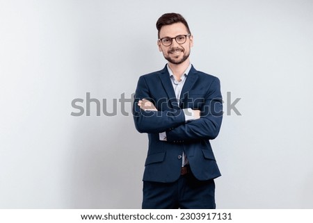 Smiling young man in a suit and glasses keeps his arms crossed. Isolated on grey background