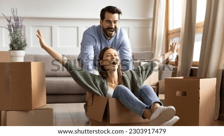 Happy loving Caucasian husband ride overjoyed millennial wife in carton box unpack on moving day to new home. Smiling young couple celebrate relocation to own house. Rental, real estate concept.