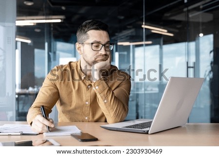 Tired and bored young man businessman, accountant, manager sitting at a table and working on a laptop and with documents. Propping his head with his hand, he looks disappointedly at the monitor. Royalty-Free Stock Photo #2303930469