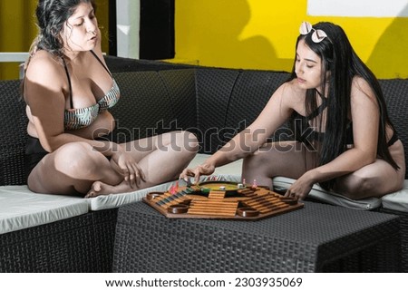Two young Latina women playing board game Parques while sitting on a black sofa and chatting happily.