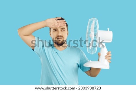 Young emaciated Caucasian man suffers from summer heat keeps fan close to him to cool off after walk or run wipes sweat with hand, dressed in casual T-shirt, stands on turquoise background Royalty-Free Stock Photo #2303938723