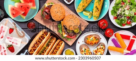 Summer BBQ food table scene. Hamburgers, hot dogs, potatoes, corn and cold treats. Top view over a dark wood banner background.